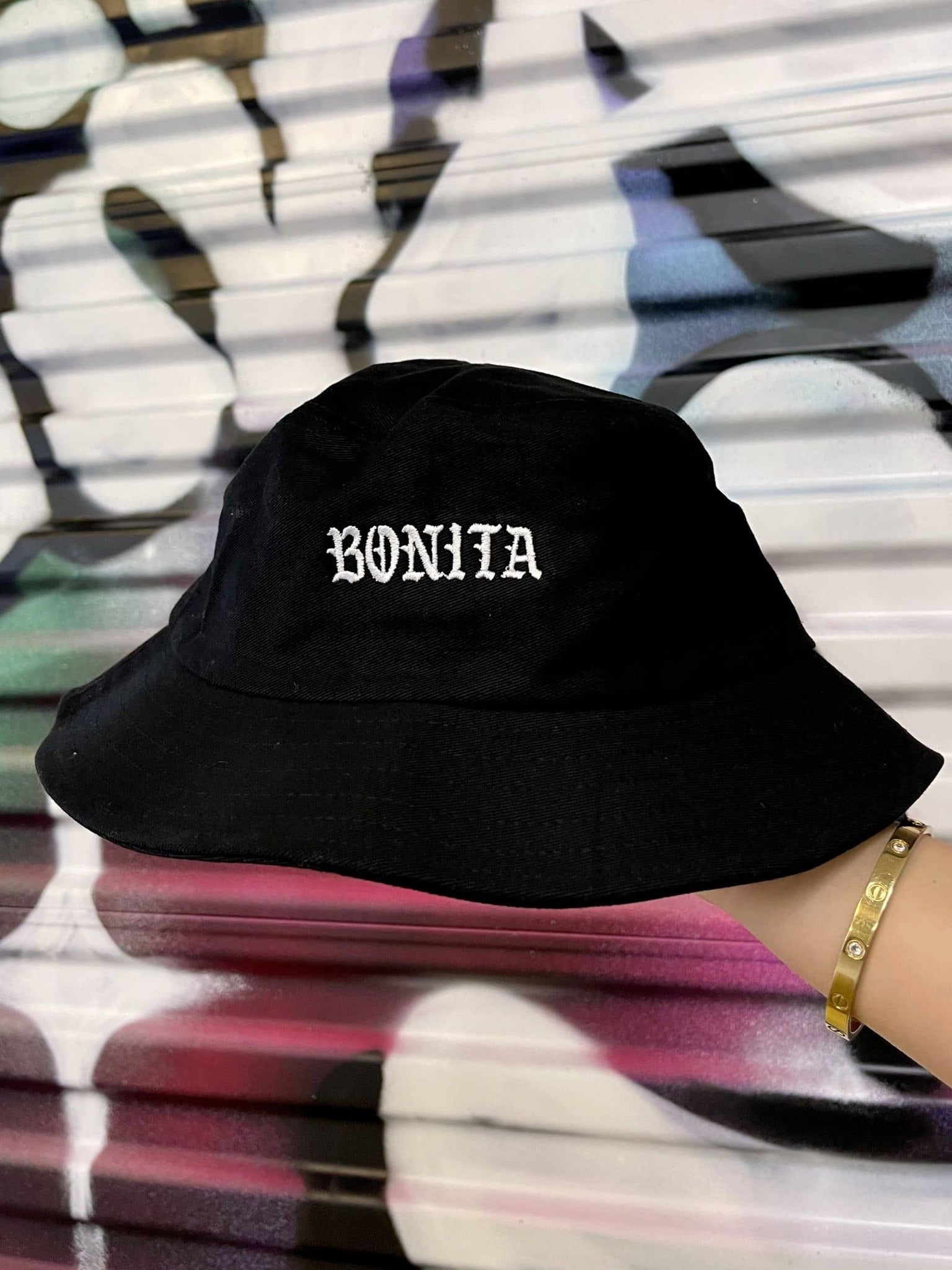 Viva La Bonita Women streetwear black Bonita Bucket hat. Hand holding black Bonita Bucket hat with graffiti background.