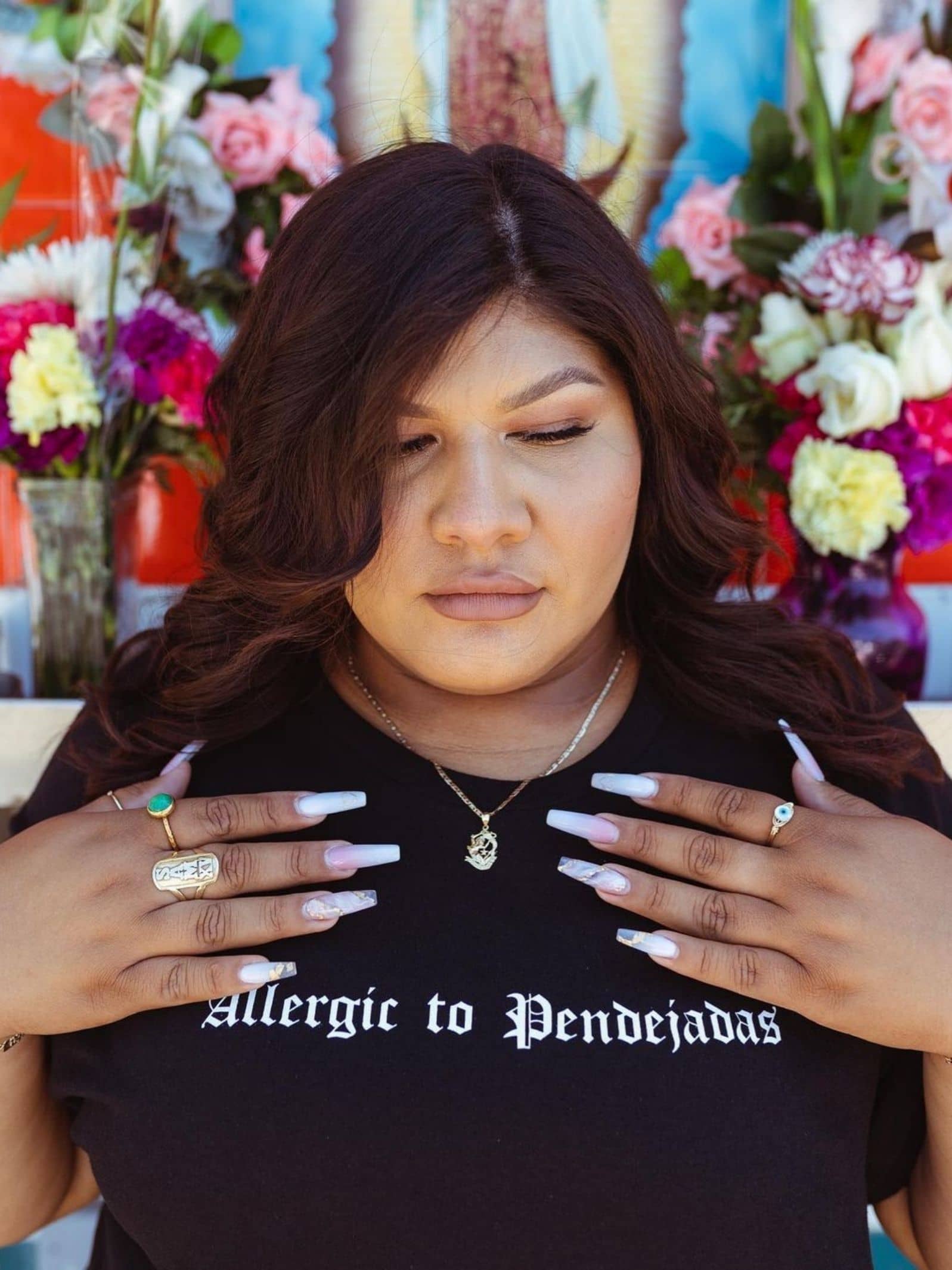 Viva La Bonita Women streetwear black t-shirt. Latina wearing black Allergic to pendejadas t-shirt with light pink stiletto nails. Flowers and virgin Mary in the background. 