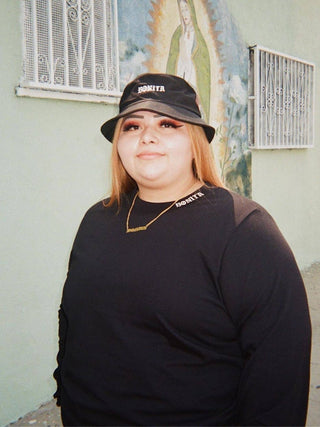 Viva La Bonita Women streetwear black Bonita Bucket hat. Model wearing black Bonita Bucket hat, gold Bonita necklace, and black mock neck long sleeve t-shirt. Model standing in front of Virgin Mary mural Los Angeles. 